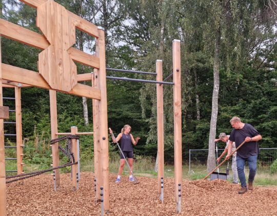 Bild på uppförande av utegym i Kågeröd. Tre personer krattar ut mjuk barkflis. man ser det träfärgade gymmets stora stälning för aktivitet. I bakgrunden björkar och lövträd.