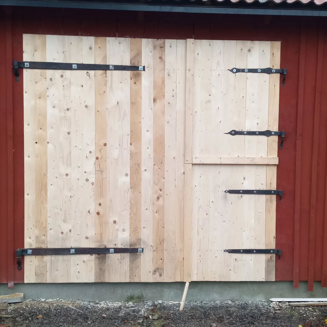 Bild på omålad traditionell ladugårdsport. I bakgrunden faluröd del av fasad på verkstad