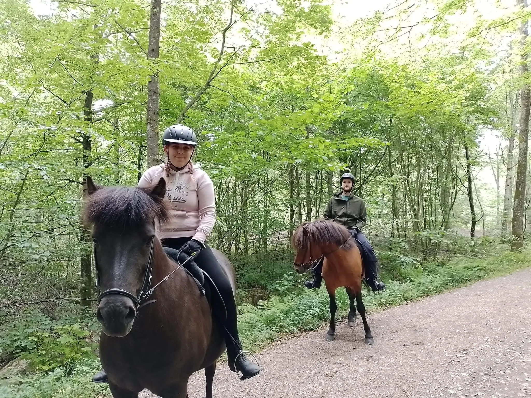 Två ryttare ute på ridtur. I bakgrunden skir lövskog och grusväg.