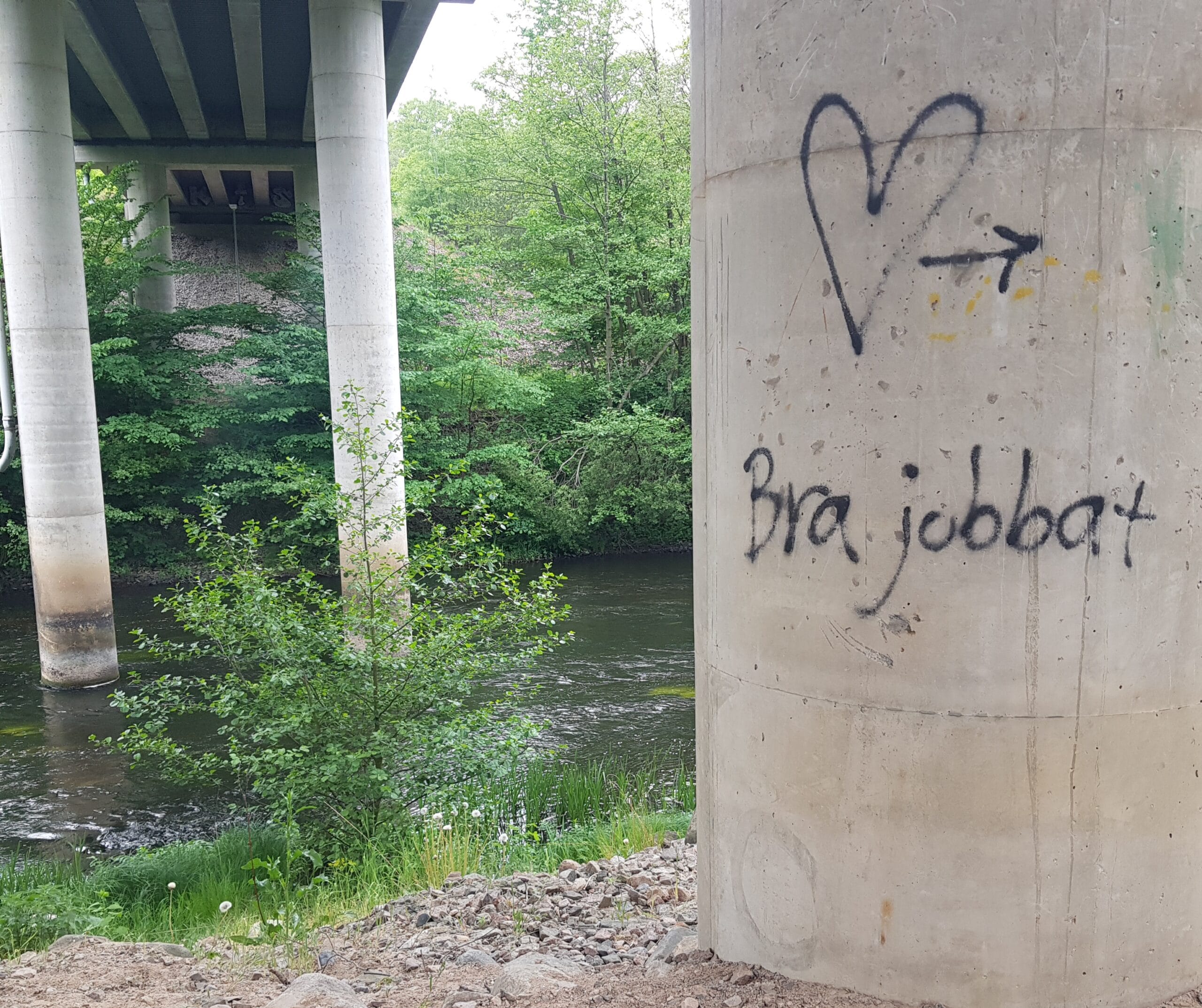 Bild på betongpelare vid å. På pelaren har någon klottrat Bra jobbat. Skir grönska