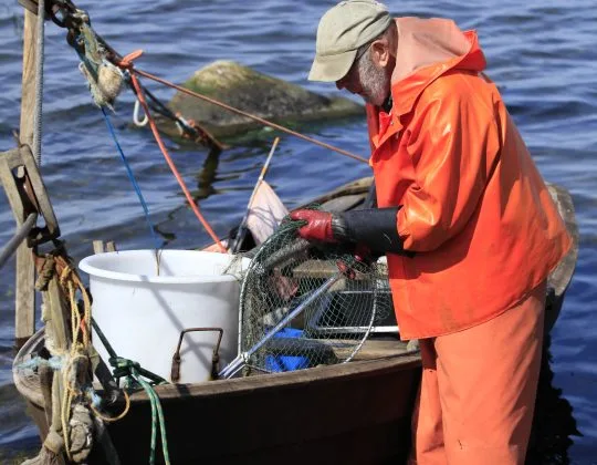 Kustfiskare vid sin båt. Havet i bakgrunden Fiskaren bär orange kläder och lägger saker i en stor vit hink