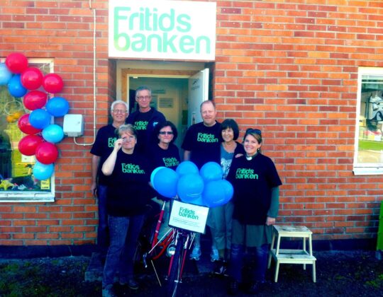Bild på engagerade personer i Fritidsbanken Sverige. PErsonerna står framför en tegelfasad med Fritidsbankens logotyp med vit bakgrund och grön text. Till vänster ser man ballonger.